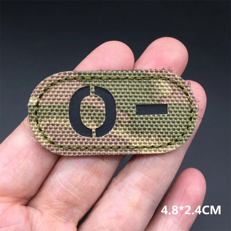 Hand holding an O- multicam IR reflective tactical blood type hook and loop patch approximately 4.8x2.4cm on a dark background
