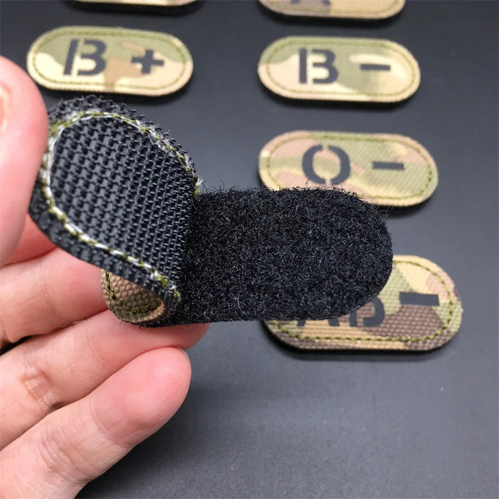 Hand holding the hook and loop backing of a multicam tactical blood type patch with other blood type patches displayed on a dark background
