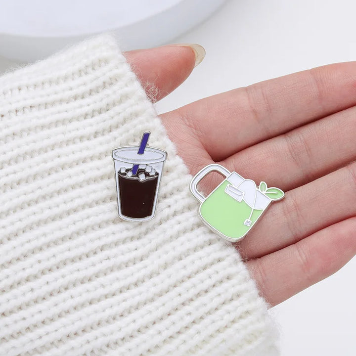 Two enamel pins shaped like a glass of iced coffee and a green mug with a leaf on a textured surface.