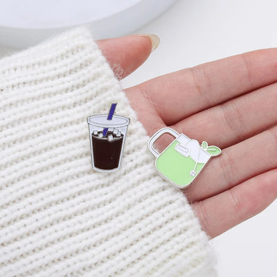 Two enamel pins shaped like a glass of iced coffee and a green mug with a leaf on a textured surface.