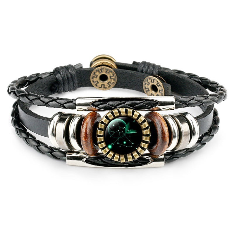 A stylish black braided and leather bracelet featuring a circular zodiac constellation center, metal accents, and wooden beads. It conveys a trendy, celestial vibe.