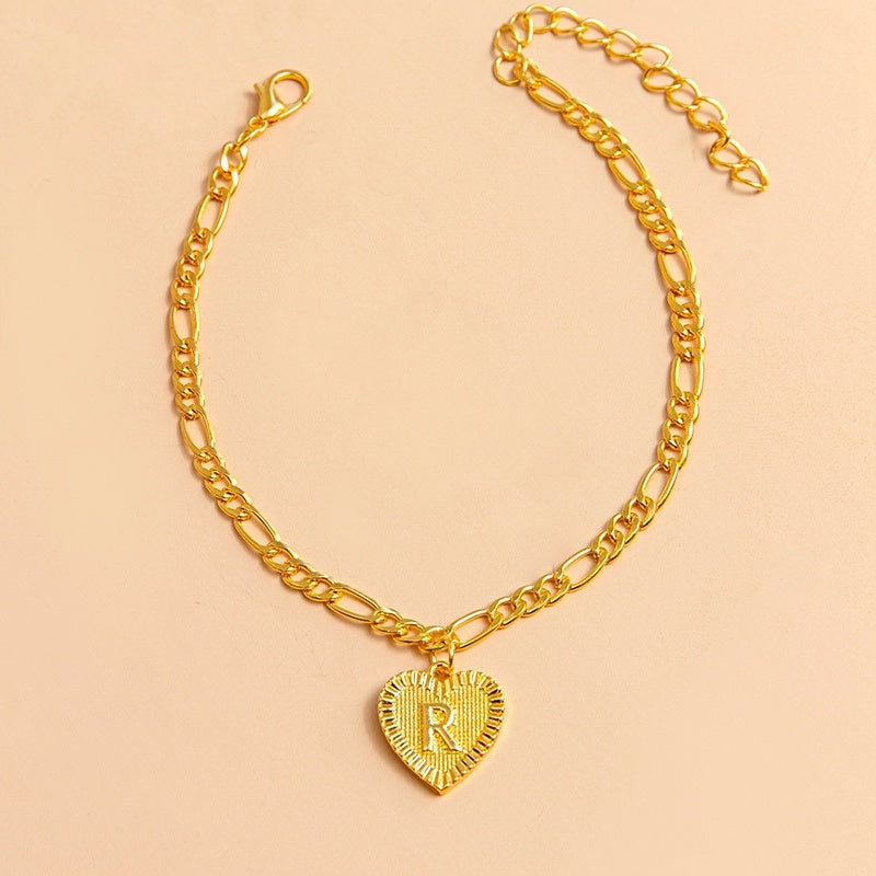 Gold chain bracelet with a heart-shaped charm featuring the letter 'R' on a beige background