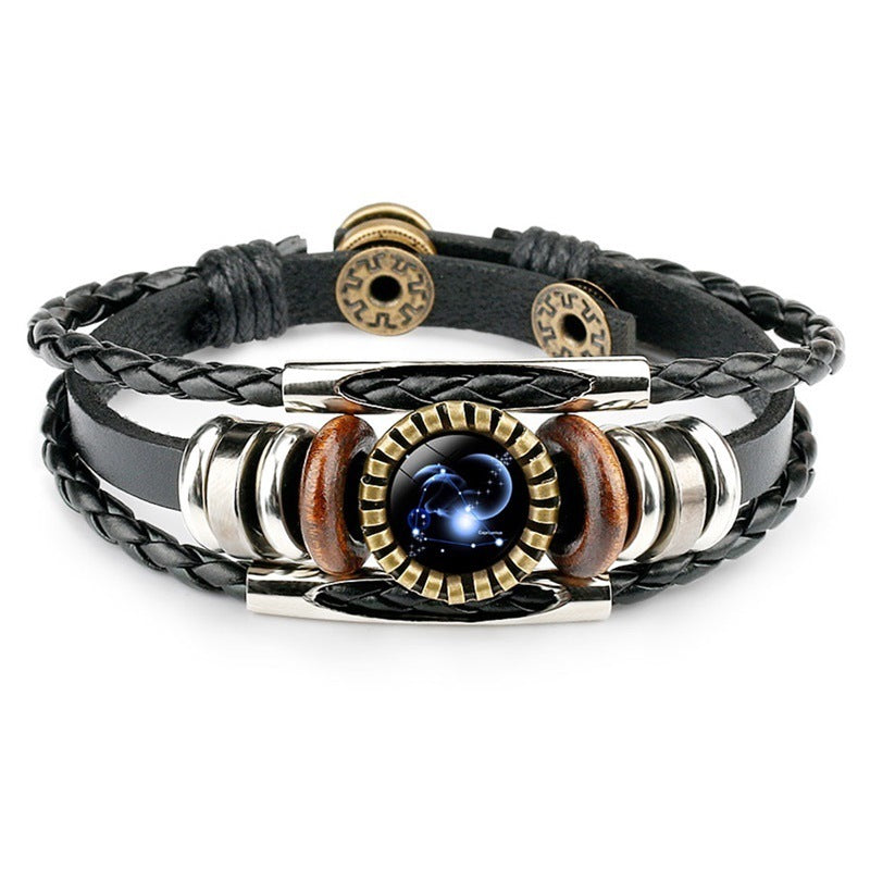 A black leather bracelet with braided and smooth bands. Features a metallic centerpiece with a celestial design, flanked by wooden and metallic accents. Elegant and modern.