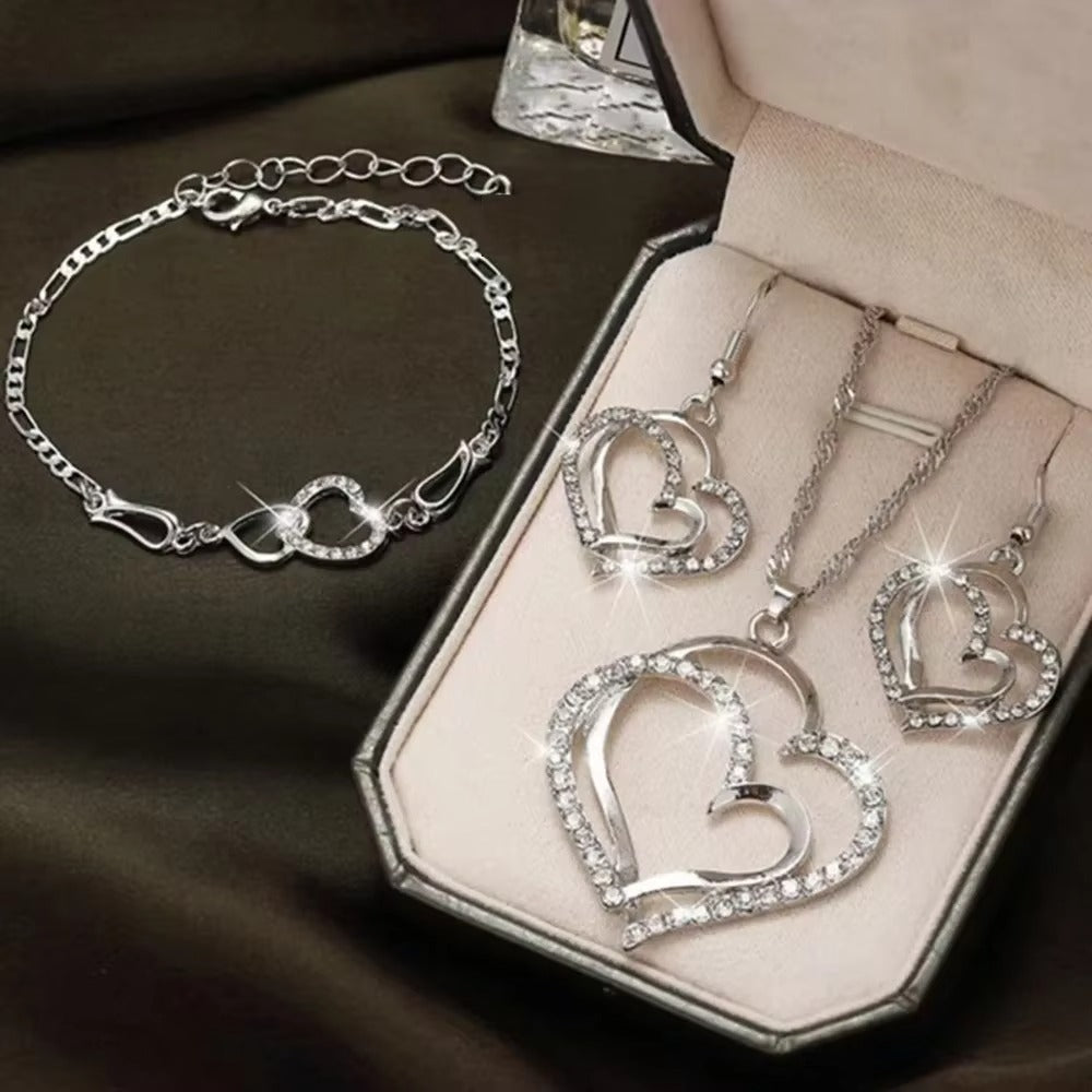 Silver heart-shaped jewelry set including necklace, earrings, and bracelet on a dark surface.